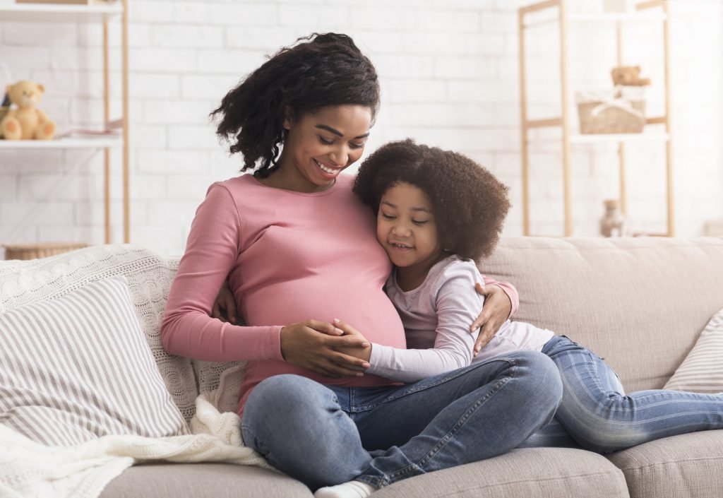 Affectionate Little Daughter Tenderly Touching Belly Of Pregnant Mom On Sofa